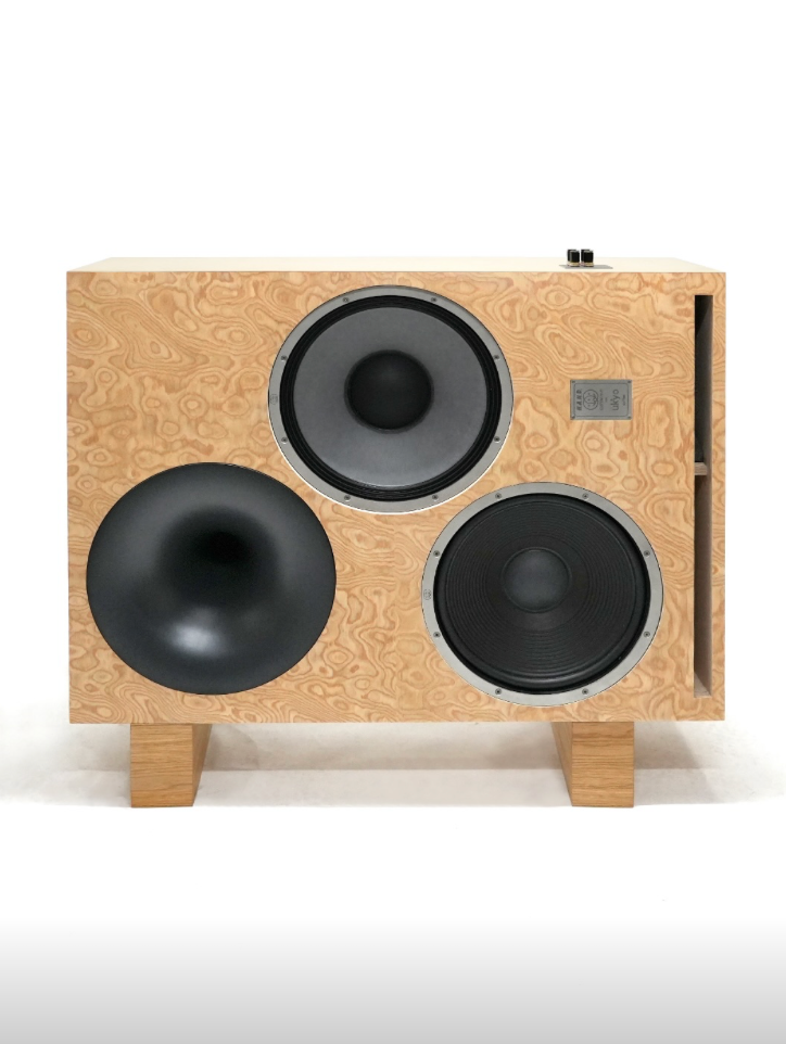 Speaker cabinet with a light natural wood grain front panel, three circular drivers (one large bass driver, two mid-range drivers), and wooden feet, shown against a plain white background.
