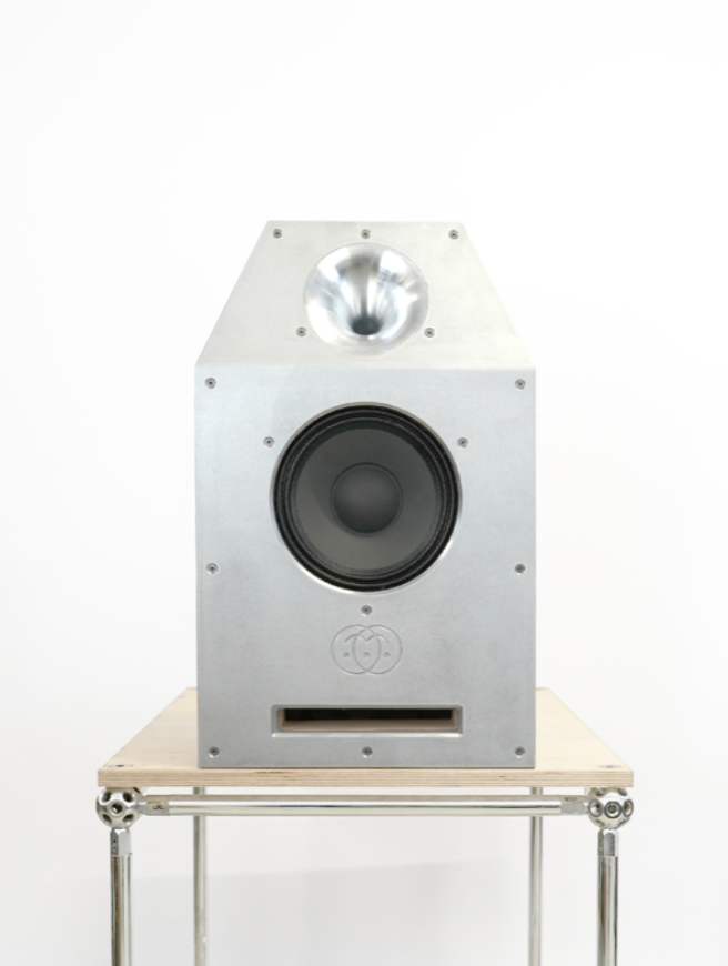 A silver aluminum tower speaker with a black woofer and a silver dome tweeter, displayed on a light wood and chrome metal stand against a white background.