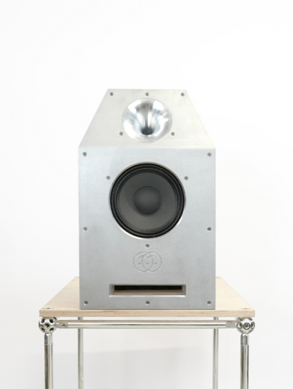 A silver aluminum tower speaker with a black woofer and a silver dome tweeter, displayed on a light wood and chrome metal stand against a white background.