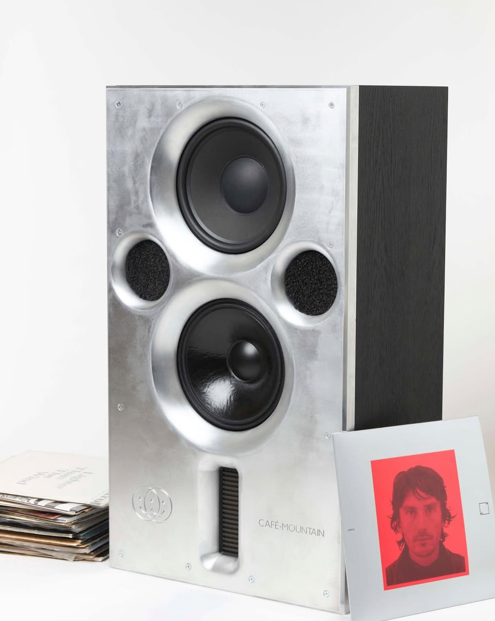 Studio monitor or bookshelf speaker with a brushed silver aluminium front baffle and a black side panel, shown next to a small printed card with a red portrait on a white background.