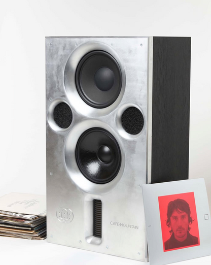 Studio monitor or bookshelf speaker with a brushed silver aluminium front baffle and a black side panel, shown next to a small printed card with a red portrait on a white background.