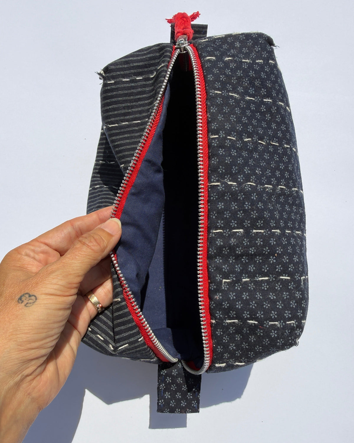 A close-up of a handmade zippered pouch with a hand holding the open zipper. The pouch has a dark blue and white patterned exterior, a red zipper track, and a navy blue interior lining.