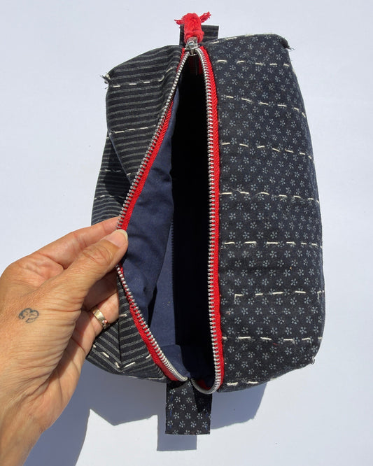 A close-up of a handmade zippered pouch with a hand holding the open zipper. The pouch has a dark blue and white patterned exterior, a red zipper track, and a navy blue interior lining.