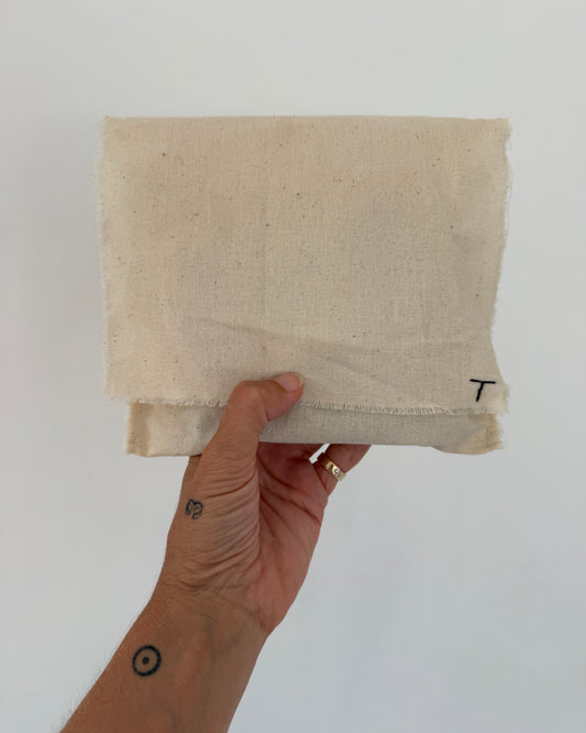 off-white woven fabric pouch with a small black letter 'T' stitched near the top edge, shown against a plain white background.